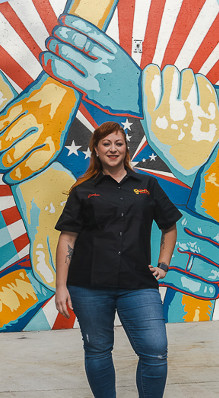 A De Anza alumna in a work uniform standing in front of a large painted mural.