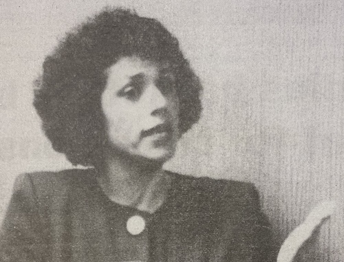 Black and white image of a woman with curly hair, wearing a dark jacket with a round pin on the collar, who is currently engaged in a speech