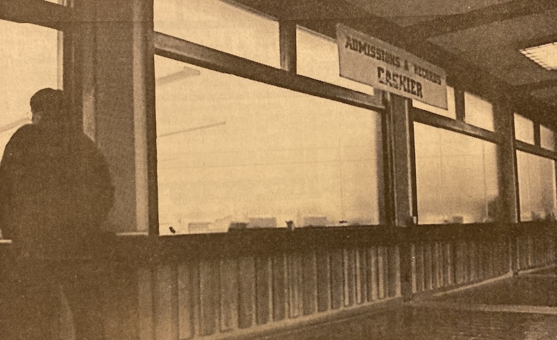 A person stands inside a building, looking out through large windows. A sign reading Admissions & Business Cashier hangs above.