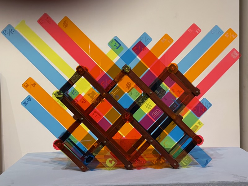 A colorful sculpture made of translucent acrylic strips in red, orange, yellow, blue, and green, woven through a wooden rack frame